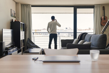 man talking on the phone looking out the window at his apartment on a workday at home