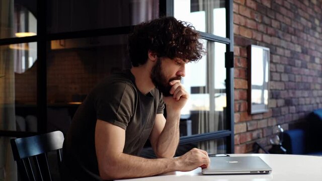 Thoughtful young man tapping his fingers on lid of closed laptop sitting at desk at home office, side view. Pensive bearded guy stuck with difficult ask, thinking on problem solution, preparing report