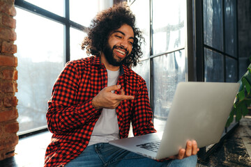 Handsome egyptian student in red plaid shirt holding a computer and pointing at it with a finger while laughing