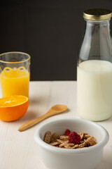 Top view of bowl with cereals, bottle of milk, half orange, glass with juice and spoon on white wooden table, black background, vertical