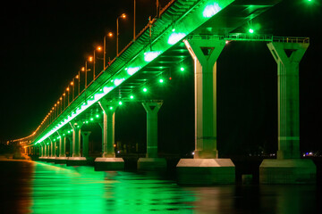 Fototapeta na wymiar Side view of colourful bridge illuminated with green color lights at the night. Bridge stands on Volga river in Russia. Multi-colored light is reflected in the water.