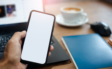 Cropped view of man hand holding blank screen smartphone with computer laptop on desk