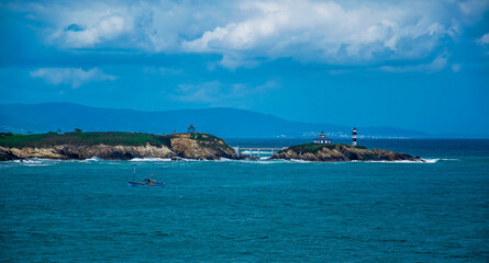 Fototapeta na wymiar Un faro en una pequeña isla unida a tierra mediante un puente, un barco pesquero de color azul, nubes de tormenta y el mar cantábrico en la costa española