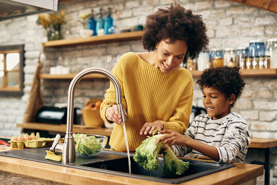 Happy black mother and son cleaning vegetables under kitchen sink.