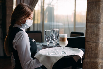 A female Waiter of European appearance in a medical mask serves Latte coffee.