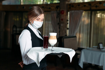 A female Waiter of European appearance in a medical mask serves Latte coffee.