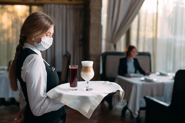 A female Waiter of European appearance in a medical mask serves Latte coffee.