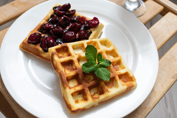 Two Belgian waffles with cherries and mint in a white plate on a wooden tray.