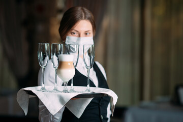 A female Waiter of European appearance in a medical mask serves Latte coffee.