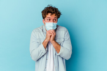 Young caucasian man wearing an antiviral mask isolated on blue background keeps hands under chin, is looking happily aside.