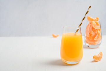 Drinking glass with straw full of sweet orange juice and tangerine or clementine segments or slices served on white wooden table against gray wall at breakfast. Image with copy space, horizontal