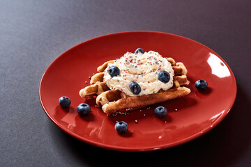 Delicious Belgian Waffle with cream and fresh blueberry on red plate on dark background