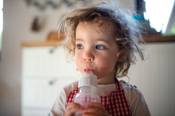 Small girl indoors in kitchen at home, drinking milk from bottle.