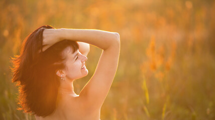 A bare-shouldered woman holds her long dark hair in her hands in a feminine way, happy in the sunset rays in a field in the fresh air. Health care, relaxation, women's health, happy woman. Copy space