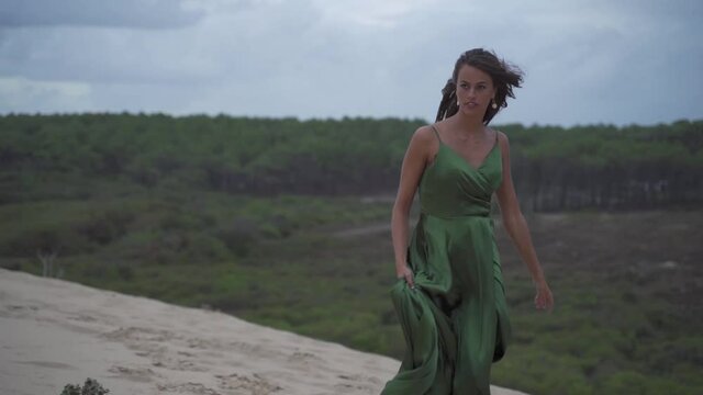 Dark haired women in a classy elegant smaragd green dress running confident at the beach - sand dune in slowmotion - cloudy sky