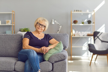 Smiling mature woman or grandmother in jeans, t-shirt and glasses sitting on sofa at home and looking at camera with room interior background. Happy elderly people, retired women concept