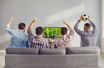 Four adult male friends with arms raised emotionally watching a football match on TV at home....