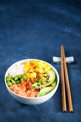 Salmon Poke bowl Raw fish salad Asian trendy food, mango, soy beans edamame, rice, avocado, served in bowl, chopsticks, blue dark background. Copyspace. Fast healthy lunch, nutrition concept.