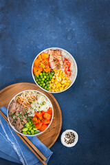 Two assorted poke bowls, flamed salmon, pulled pork, vegetables, rice, sauces. Top view, close-up. Hawaiian dish, blue dark background. Trendy asian food. Healthy and clean eating concept.