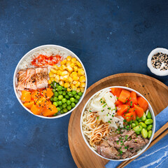 Two assorted poke bowls, flamed salmon, pulled pork, vegetables, rice, sauces. Top view, close-up. Hawaiian dish, blue dark background. Trendy asian food. Healthy and clean eating concept.