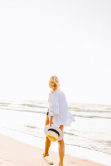 Beautiful blonde slim woman wearing black bikini and white shirt walk on beach of sea or ocean against the sun. Youth, vacation, travel, fashion underwear