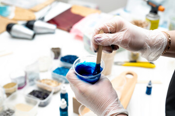 artist's hands in disposable transparent gloves, stirring a blue dye with a wooden spatula in a...