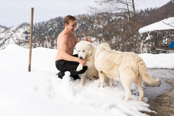 A man with a naked torso caresses a white dog in the snow