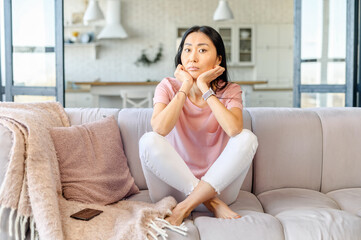 Young adult Asian mixed-race female domestic housewife with long black hair sitting on the couch, feeling bored and demotivated, does not want to do tasks, upset with the message from phone, unhappy