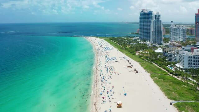 Top view of crowded tropical Atlantic ocean beach showing colorful beach umbrellas and people relaxing on sunny summer day. Drone water shot with perfect teal green blue shore. Miami beach, USA 4K