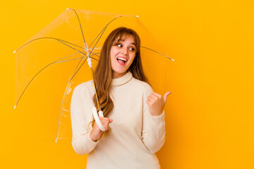 Young caucasian woman holding a umbrella isolated points with thumb finger away, laughing and...