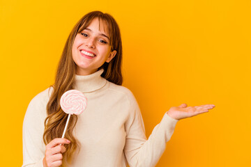 Young caucasian woman holding a lollipop isolated on yellow background showing a copy space on a palm and holding another hand on waist.