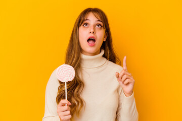 Young caucasian woman holding a lollipop isolated on yellow background pointing upside with opened mouth.