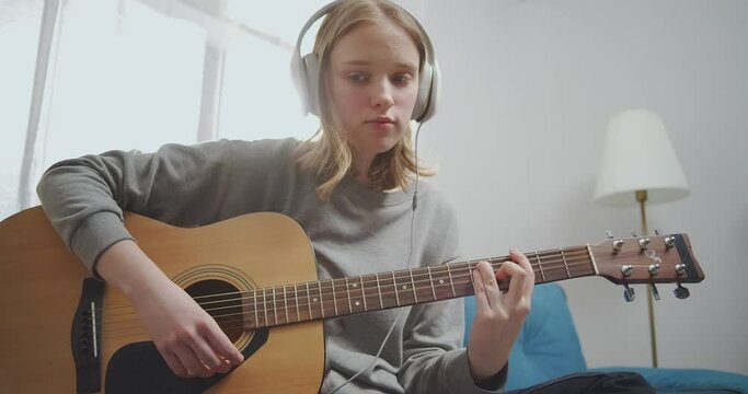Happy young girl picks up a guitar chord with her fingers with her left hand. A woman with headphones listens to music.