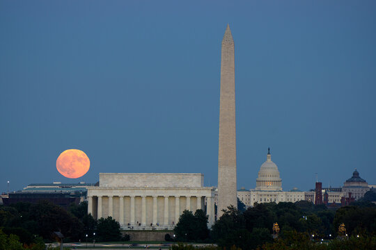 Large full moon rises through the haze over the Capitol building in Washington DC with Lincoln Memorial and Washington Monument aligned.