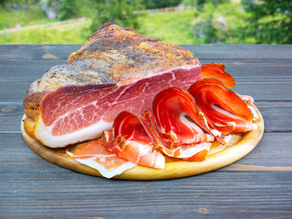 Whole speck with slices. Typical South Tyrolean raw ham outdoors. Italian food. Table in nature, in...
