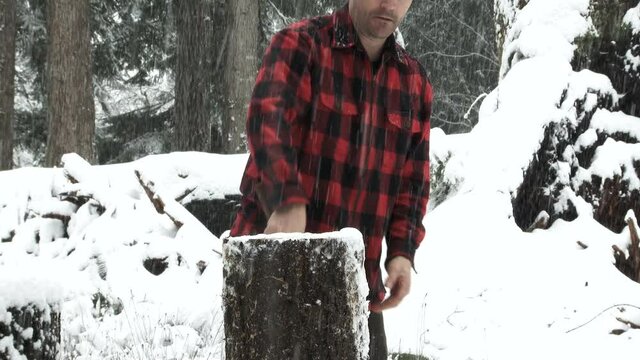 Lumberjack out in snowy winter forest takes a hard swing at a log, splitting it on first try, slow motion.