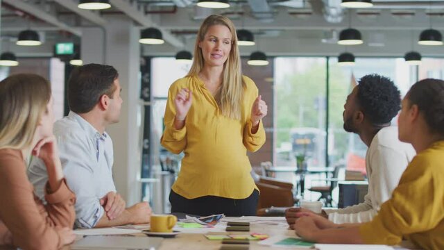 Pregnant businesswoman standing and giving presentation on performance to colleagues sitting around table in modern open plan office - shot in slow motion