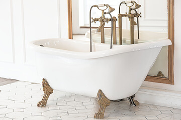 A beautiful white Baroque bathtub with gold legs and a faucet stands in a bright, spacious room next to a large mirror. The concept of a vintage interior of a house or photo studio.