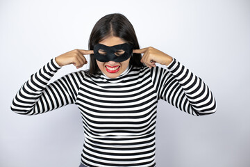 Young beautiful brunette burglar woman wearing mask covering ears with fingers with annoyed expression for the noise of loud music. Deaf concept.