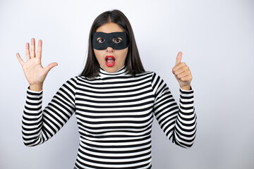 Young beautiful brunette burglar woman wearing mask showing and pointing up with fingers number six smiling confident and happy