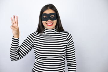 Young beautiful brunette burglar woman wearing mask showing and pointing up with fingers number three smiling confident and happy