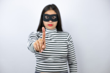 Young beautiful brunette burglar woman wearing mask showing and pointing up with fingers number one while is serious