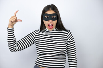 Young beautiful brunette burglar woman wearing mask smiling and confident gesturing with hand doing small size sign with fingers . Measure concept.