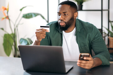 Concentrated successful stylish african american man sits at his desk at home or office, uses mobile phone and bank card to pay bills online or shopping online