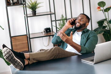 Relaxed confident african american male freelancer or business man sitting at work desk in the office with legs on table and talking on the cellphone with business partner, gesturing hand, smiling