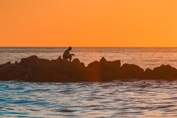 The silhouette of a lonely man sitting in meditation on the rocks of a breakwater near the sea coast against the orange sunset.