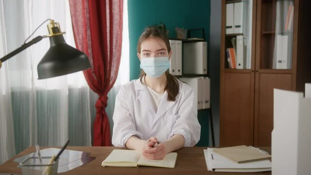 Medical Student In Mask Is Using Online Video Conference For Remote Learning. Nurse Is Studying At Home. Doctor Is Working Remotely. X-rays, Notes And Books At The Table. Woman Is Looking At Camera. 