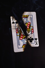A charred and burnt playing card, the king of clubs, split apart diagonally with an abstract smoke pattern in the background.