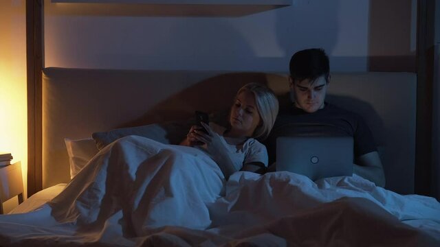 Gadgets night. Digital couple. Modern technology. Home leisure. Woman using mobile phone in bed while husband busy working online on laptop late in dark bedroom with blue warm yellow lamp light.