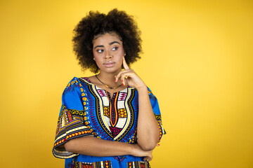 African american woman wearing african clothing over yellow background serious face thinking about question with hand on chin, thoughtful about confusing idea